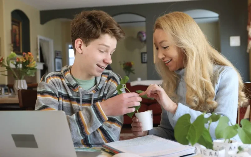 Un niño pequeño y una mujer examinan juntos con alegría una pequeña planta en una mesa. La acogedora habitación tiene una iluminación cálida, flores al fondo y un ordenador portátil abierto.