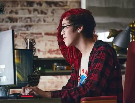 Una persona con el pelo largo y rojo y gafas está concentrada frente a un ordenador en una acogedora habitación con paredes de ladrillo. Lleva una camisa a cuadros, lo que transmite concentración.