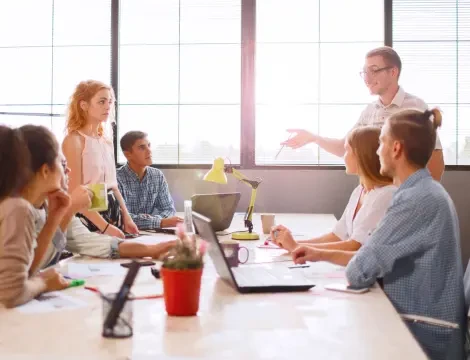 Grupo de seis personas en una oficina luminosa con grandes ventanas, conversando alrededor de una mesa con ordenadores portátiles y documentos. Una persona está de pie, hablando animadamente.