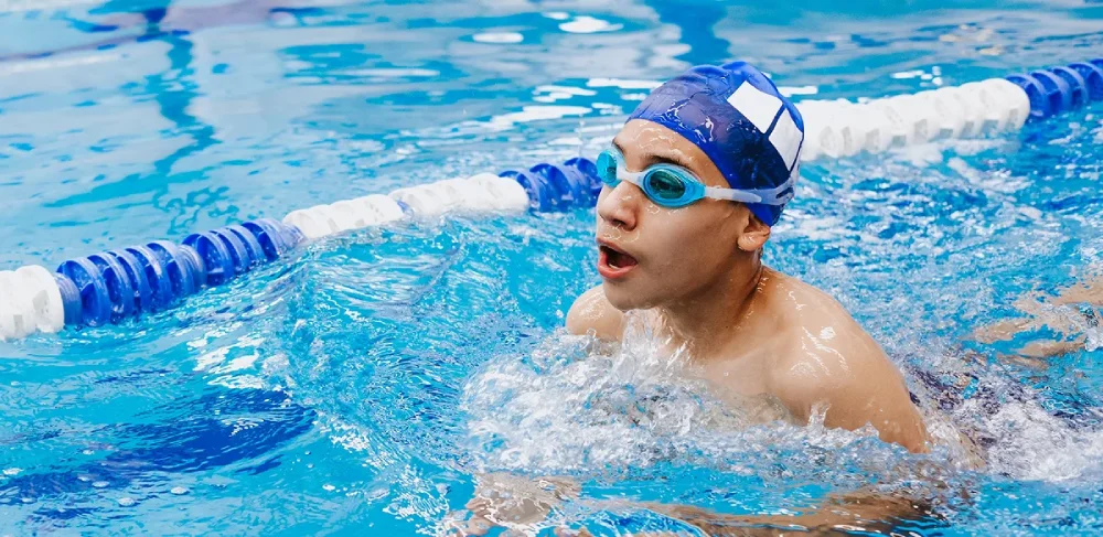 Joven nadador con gorro azul y gafas de natación haciendo braza en la piscina, con salpicaduras de agua a su alrededor. Se ven los separadores de carriles, lo que transmite concentración y energía.