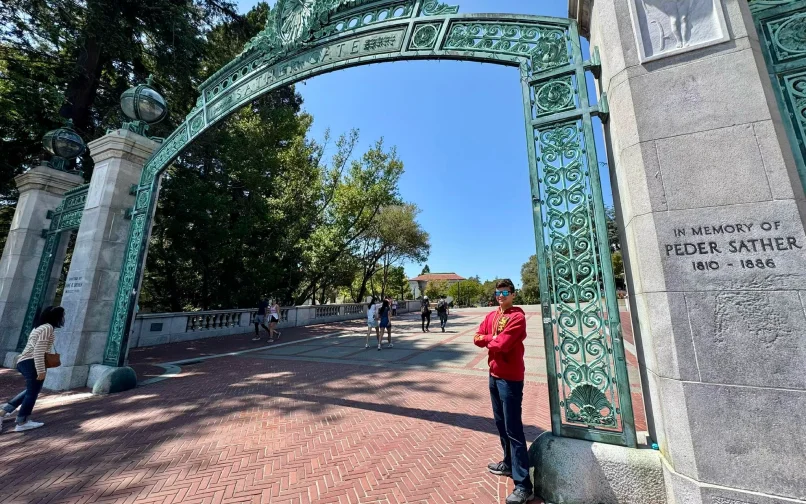 Una persona de pie junto a la base de un gran arco decorativo en el campus de una universidad. El arco, de metal verde y ornamentado, cruza una amplia pasarela por la que caminan varias personas. Al fondo se divisan árboles altos y edificios del campus, y en un pilar de piedra junto al arco se lee la inscripción: «En memoria de Peder Sather, 1810-1886».